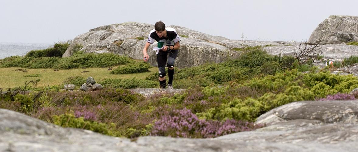 Löpare som läser kartan i ljungen, med en klippa och havet i bakgrunden